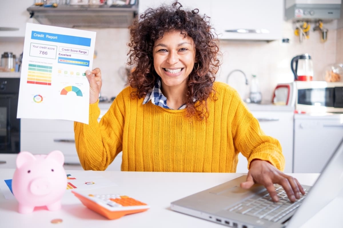 Someone holding up a document labeled credit score report next to a piggy bank, calculator, and laptop